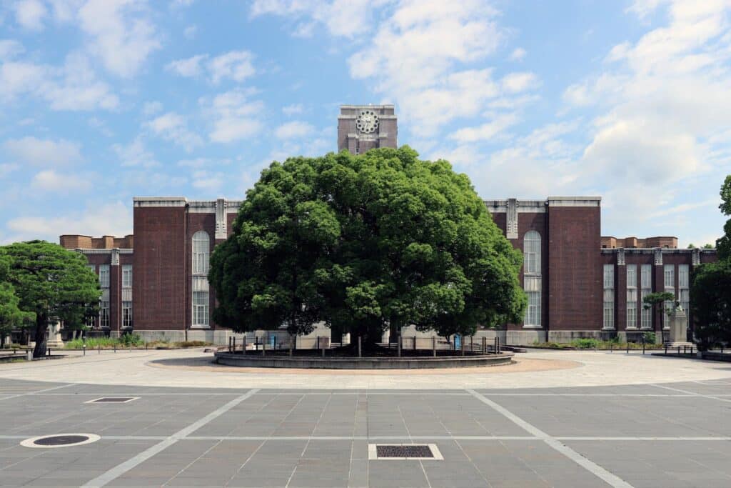 Photograph of a large, leafy tree centered in front of a historic brick building with a clock tower under a partly cloudy sky.