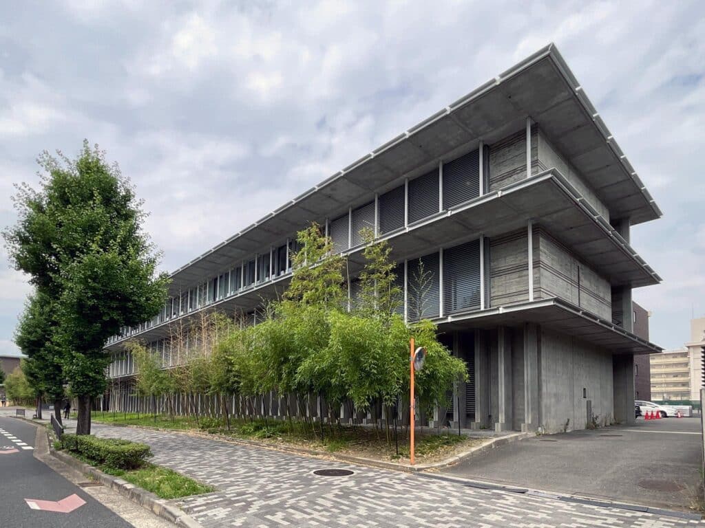 Photograph of a modern, multi-story concrete building with horizontal louvers and large overhanging roofs, surrounded by trees and a paved walkway. The structure features a minimalist design with vertical columns and greenery lining one side, set against a cloudy sky.