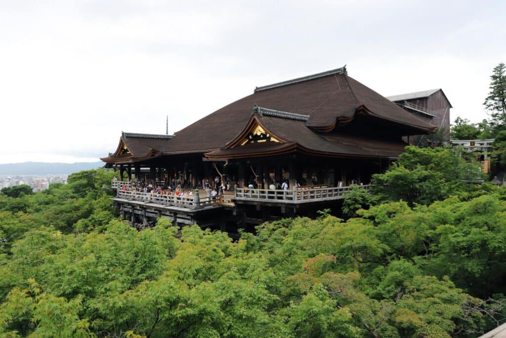 Photograph of a large traditional Japanese wooden temple surrounded by dense green foliage, with numerous visitors standing on the elevated veranda. The temple features dark brown curved roofs with gold accents, showcasing classic architectural design and providing a scenic view over the lush forest.