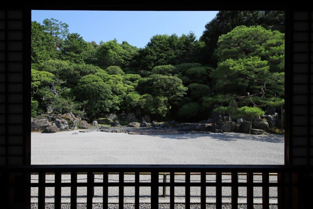 Photograph of a traditional Japanese rock garden framed by a wooden veranda railing and window. The garden features meticulously raked white gravel in the foreground and dense green shrubs and trees with rocks in the background, showcasing a serene and minimalist landscape design.