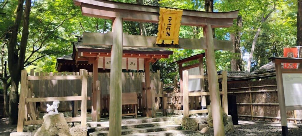 Photograph of a traditional Japanese Shinto shrine entrance featuring a wooden torii gate with a yellow hanging plaque displaying Japanese characters. Surrounding elements include wooden fences, stone steps, and lush green trees providing a serene and natural setting.