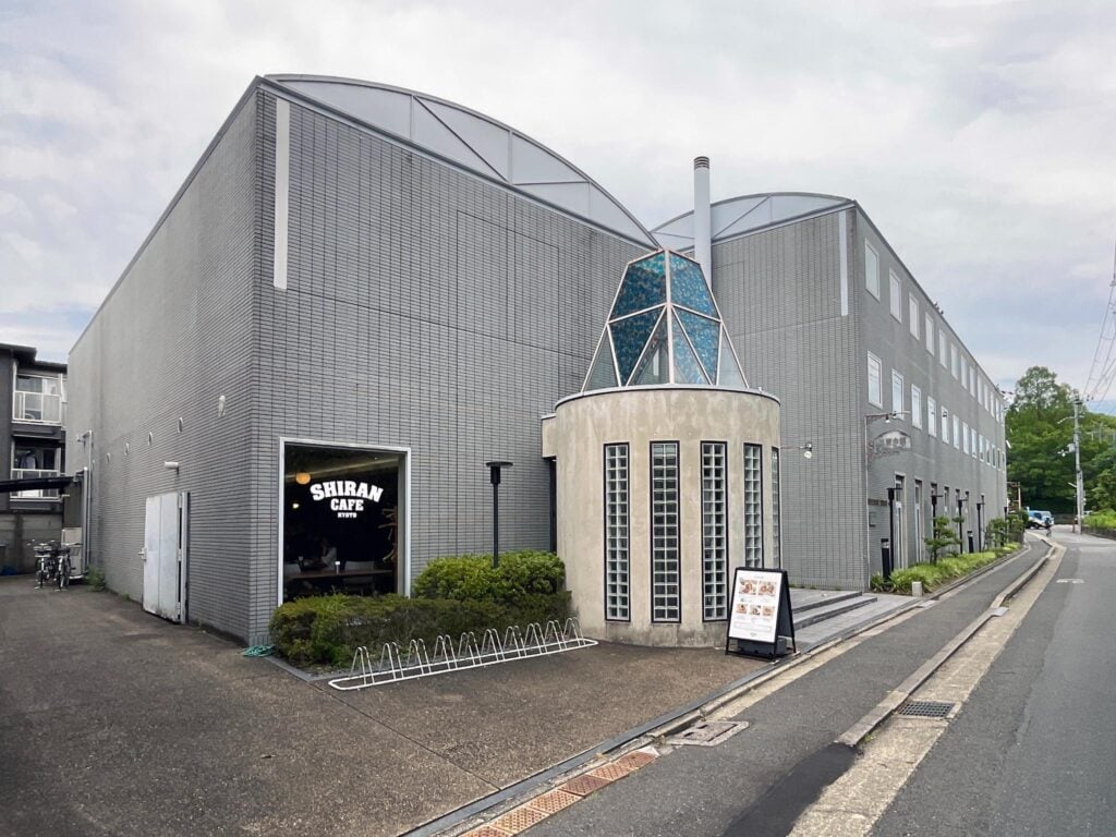 Photograph of a modern, gray brick building housing Shiran Cafe, featuring a unique cylindrical glass block structure with a blue-tinted glass pyramid on top near the entrance. The scene includes a sidewalk, bike rack, and signage displaying cafe information, set against an overcast sky.