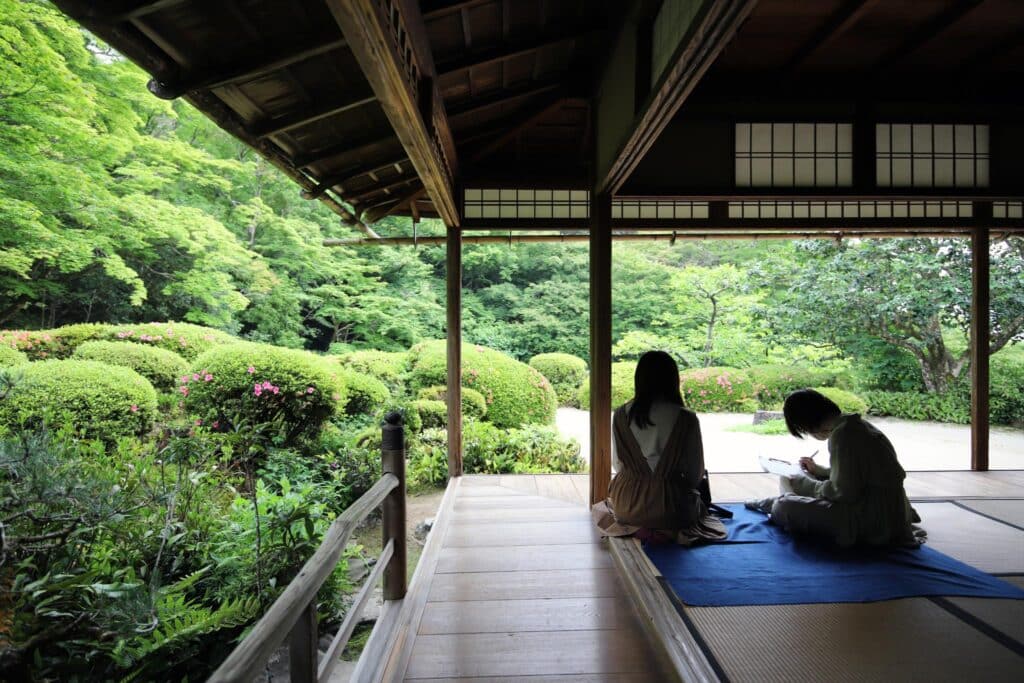 Photograph of two people sitting on a blue mat inside a traditional Japanese wooden pavilion overlooking a lush green garden. The scene highlights serene natural surroundings with manicured bushes, trees, and soft natural light filtering through open sliding doors.