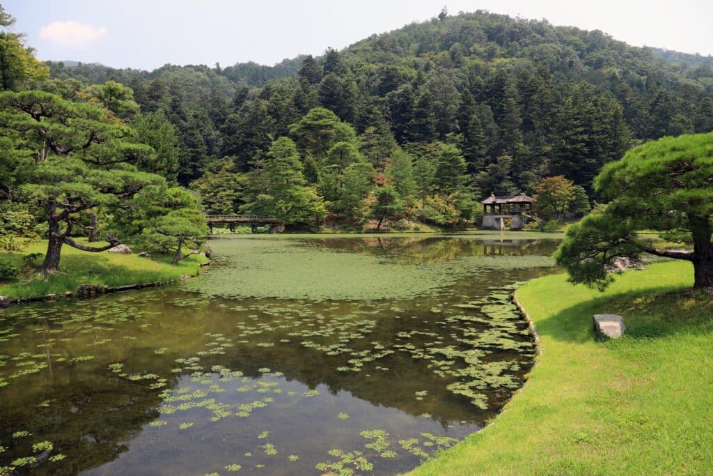 Photograph of a serene Japanese garden featuring a pond with floating lily pads, surrounded by manicured trees and lush greenery. A small traditional wooden pavilion is visible near the pond's edge, with forested hills in the background under a partly cloudy sky